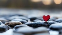 Small red heart symbol placed on smooth river stones with soft bokeh background creating romantic peaceful atmosphere for wellness meditation concepts.