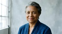 Confident mature African American businesswoman in blue blazer smiling warmly in modern office setting with natural window light for corporate portraits.