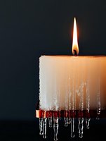Burning white candle with frozen icicles hanging from melted wax against dark background, creating dramatic contrast between warm flame and cold ice formations.