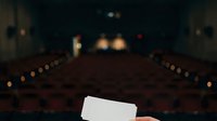 Blank movie ticket held in focus with blurred cinema auditorium seating and screen in background, suitable for entertainment promotions and ticket sales marketing.