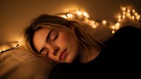 Young woman sleeping peacefully on pillow with warm glowing fairy lights creating magical bokeh effect in dark bedroom, long hair spread on white bedding.