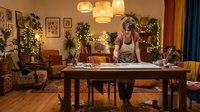 Young blonde woman cooking in festive kitchen decorated with Christmas lights and holiday garland, preparing meal at wooden table in cozy home interior.