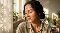 Middle-aged woman with dark wavy hair and olive complexion, wearing casual beige shirt, sitting indoors near window with plants, looking down thoughtfully in natural daylight.