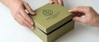Two hands holding olive green kraft box with leaf logo on white background, suitable for packaging mockups, branding presentations, and product design templates.