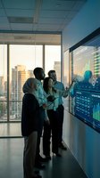 Diverse business team analyzing financial data on interactive digital display in modern office with city skyline view during golden hour sunset.