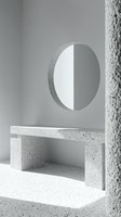 Minimalist architectural interior with terrazzo bench and round mirror on gray wall. Strong shadows create geometric shapes in monochromatic white space.