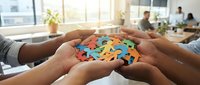 Multiple hands holding colorful puzzle pieces together in office setting, representing teamwork, collaboration, problem-solving and unity in business environment.