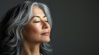 Serene mature Asian woman with flowing silver gray hair and closed eyes in peaceful meditation pose against dark background, radiating calm and wellness.