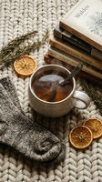 Cozy hygge flatlay with steaming tea mug, stacked books, dried orange slices, wool socks, and rosemary on chunky knit blanket.