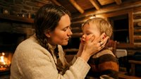 Caucasian mother gently holds toddler son's face in rustic log cabin with warm fireplace glow, tender family moment capturing love and care.