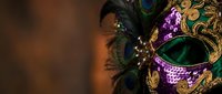 Ornate Mardi Gras carnival mask with peacock feathers, gold filigree details and purple sequins against warm bokeh background for festive celebrations.
