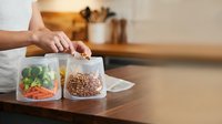 Person's hand selecting between two clear storage containers with fresh vegetables and grains on kitchen counter for meal prep and healthy eating organization.