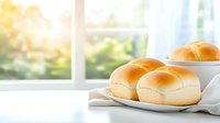 Fresh baked dinner rolls on white plate with natural window lighting and soft morning glow, homemade bread served on kitchen table with linen napkin.