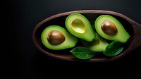 Fresh halved avocados with pits and green leaves arranged in dark wooden bowl on black background, dramatic food photography styling.