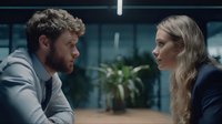 Young Caucasian man and woman in business attire having serious conversation in modern office with glass walls and plants in background.