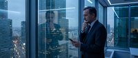 Professional businessman in dark suit talking on phone while holding tablet in modern glass office overlooking city skyline at dusk with urban lights.