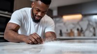 African American male chef sprinkling flour on kitchen counter while preparing dough, wearing white t-shirt, focused on culinary work in modern kitchen setting.