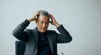 Middle-aged man with gray hair and Asian ethnicity wearing dark business suit, hands to head in stressed or thoughtful pose against white background, suitable for corporate, business, and professional concepts.