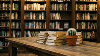 Stacked books and potted cactus on wooden table in front of library shelves, creating a scholarly and botanical study environment for educational and creative projects.