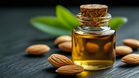 Golden almond oil in glass bottle with cork stopper surrounded by raw almonds and green leaves on dark wooden surface for natural skincare and aromatherapy.