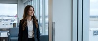Professional businesswoman in navy blazer standing by airport terminal windows looking optimistic during business travel or corporate trip.