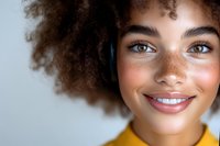 Young African American woman with natural curly hair and freckles smiling warmly at camera, wearing yellow top against light background. Close up portrait shows radiant skin.