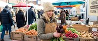 Young blonde woman in beige knit hat and brown apron smiling while holding fresh vegetables at bustling outdoor farmers market with wooden crates full of produce.