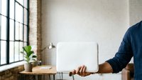 Young man with medium skin tone in casual business attire holding laptop computer in modern office workspace with natural light and minimalist decor for technology and business concepts.