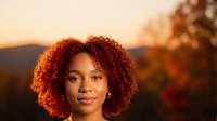 Young African American woman with curly hair smiling outdoors during golden hour sunset with warm lighting and blurred natural background.