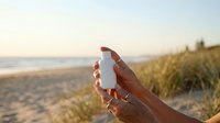 Sunscreen bottle held in hand at beach during golden hour with ocean and dunes in background for skincare and sun protection advertising.
