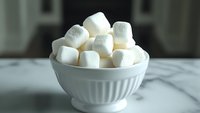White fluffy marshmallows piled in ceramic ribbed bowl on marble surface, soft natural lighting creates gentle shadows highlighting sweet treat texture.