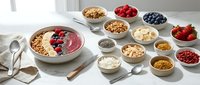 Overhead view of breakfast bowls with granola, yogurt, berries, nuts, and toppings arranged on white surface for healthy eating and meal prep content.