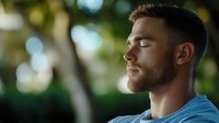 Young Caucasian man with beard in blue t-shirt meditating outdoors with eyes closed, peaceful expression, natural bokeh background with sunlight and greenery.
