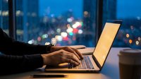 Professional working late on laptop computer in modern office with city lights bokeh background during evening hours for business productivity.