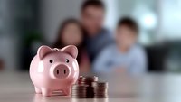 Pink ceramic piggy bank with stacked coins on wooden table, blurred family background represents financial planning and household savings concept.