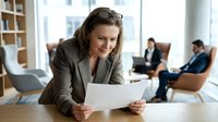 Mature woman with brown hair and light complexion, wearing professional beige blazer, holding document at wooden desk in modern office with colleagues in background.