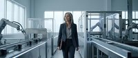A professional woman in business attire walks through a modern laboratory or manufacturing facility with advanced robotic equipment and machinery on both sides.