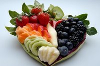 Heart-shaped fruit platter with fresh strawberries, blackberries, blueberries, kiwi, banana, and melon slices arranged artistically on white background.