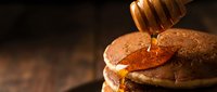 Golden honey dripping from wooden dipper onto stack of fluffy pancakes against dark rustic background for breakfast and cooking concepts.