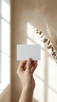 Female hand holding blank white business card mockup with natural sunlight shadows and dried eucalyptus pampas grass in warm beige interior for branding.