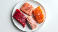 Fresh raw meat and fish pieces on white plate including beef steak, salmon fillet, and tuna. Top view of protein-rich ingredients for healthy cooking.