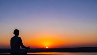 Silhouette of person meditating in lotus position during golden hour sunset over calm water with colorful sky gradient for wellness and mindfulness concepts.