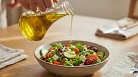 Young woman pouring golden olive oil over fresh vegetable salad in bowl for healthy eating and cooking preparation.