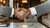 Professional business handshake between two people wearing formal suits against blurred office background with warm lighting, symbolizing partnership and agreement.