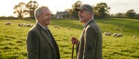 Two senior Caucasian men in tweed jackets chat warmly in a green pasture with sheep grazing at golden hour on a rural farm.