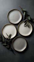 Four empty ceramic plates with gold rims arranged on dark gray surface with fresh eucalyptus branches. Overhead flat lay composition for tableware, dining, and home decor design projects.