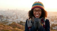 Happy young African American woman in colorful knit hat and glasses smiling warmly at camera during golden hour with city skyline background.