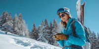 Female skier in blue jacket using smartphone on snowy mountain slope with ski equipment and snow-covered pine trees against clear blue sky.