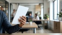 Man in business casual attire holding blank white tablet device in modern office environment with colleagues working at desks in background for corporate technology and business applications.