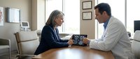 Middle-aged Caucasian woman patient consulting with Hispanic male doctor in modern medical office discussing brain scan results on tablet device.